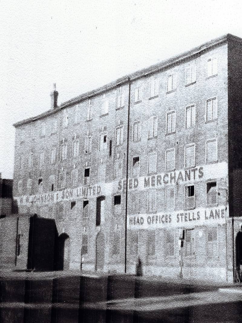 A black and white photograph of Johnson Seed Factory. It is a five storey brick building. Johnson and Son Seed Merchants, Head Offices Steel Lane is painted on the front in white.