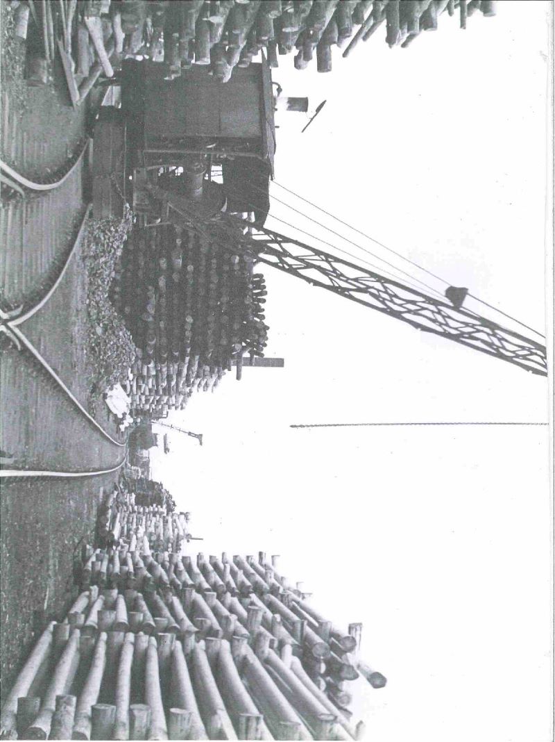 A black and white photograph of the wood yard and the railway crane. Wood is stacked throughout the yard. There is a railway crane in the foreground, and another crane futher down the track that leads down the middle of yard.