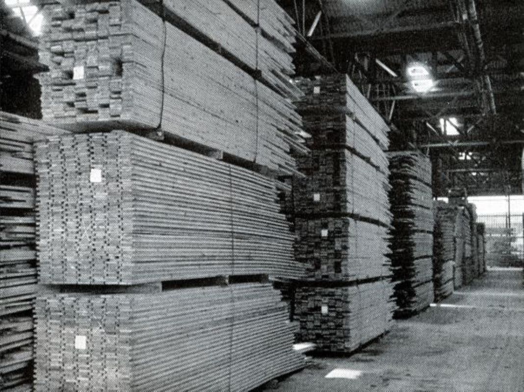 A black and white photograph of wood planks in storage. Stacks of planks are lashed together in blocks and stacked on top of one another. The wood is being stored in a large warehouse.