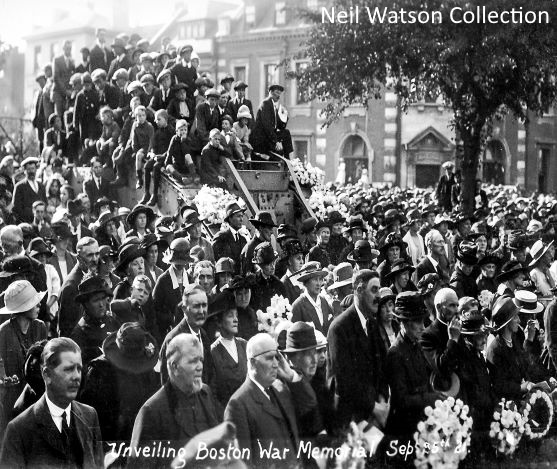 A black and white photograph of the unveiling of the Boston war memorial in 1921. There is a large crowd of people stood in the town centre, with many holding wreaths. A group of children are stood on top of a cart to see over the crowd.