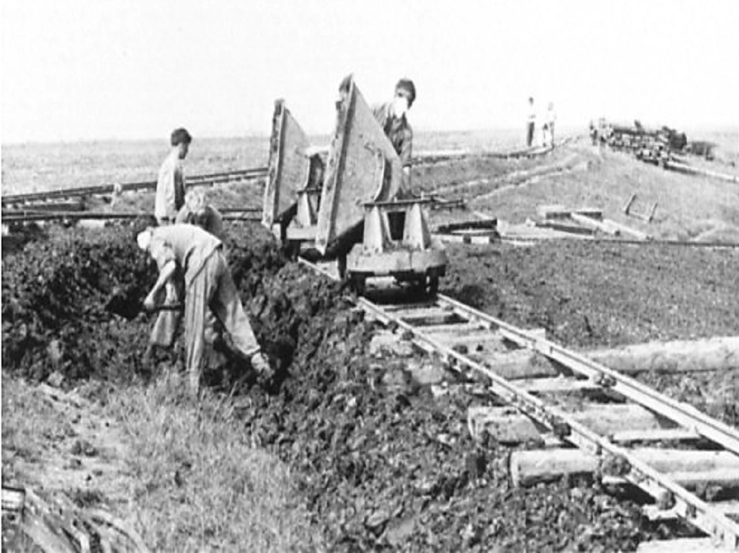 A black and white photograph of Borstal Boys from the north sea camp at work building the sea bank. Barrows of earth, carried along a track, are tipped and emptied while more men shovel to build up the bank.