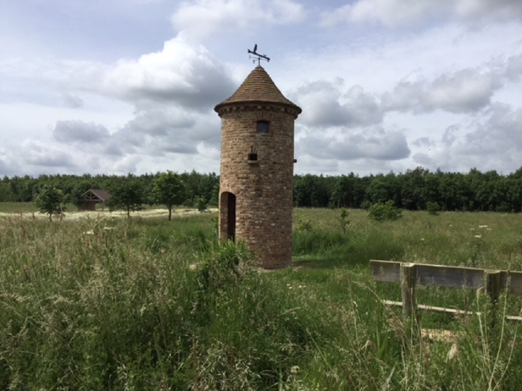 A photograph of an owl tower. It shows a small, round, brick tower with a steep tile roof. A large doorway is in one side of the tower. A wind vane is on top of the roof. 