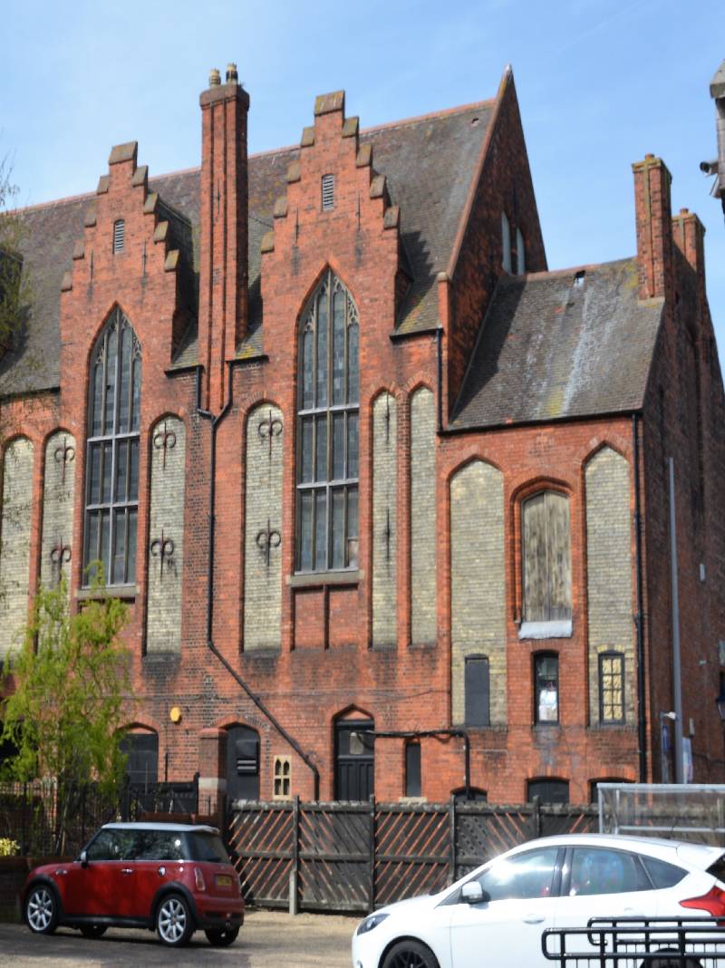 A coloured photograph of the rear of Shodfriars Hall. It is a large red brick building with tall and slender arched windows and a steep roof. The widows are framed in a stepped design. There are several very tall chimneys.